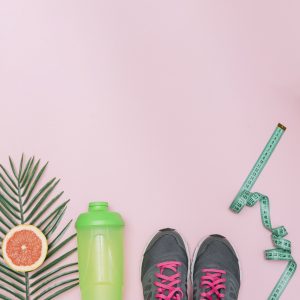 Pedaço de laranja, copo de água, tênis e fita métrica lado a lado em fundo rosa, representando o que é necessário para perder peso.
