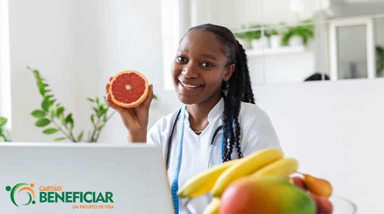 Nutricionista negra, com o cabelo trançado e usando um jaleco branco segura uma laranja. Em primeiro plano, estão algumas frutas indicadas para um emagrecimento saudável, como banana e manga, desfocadas. Ela está na frente de um notebook.