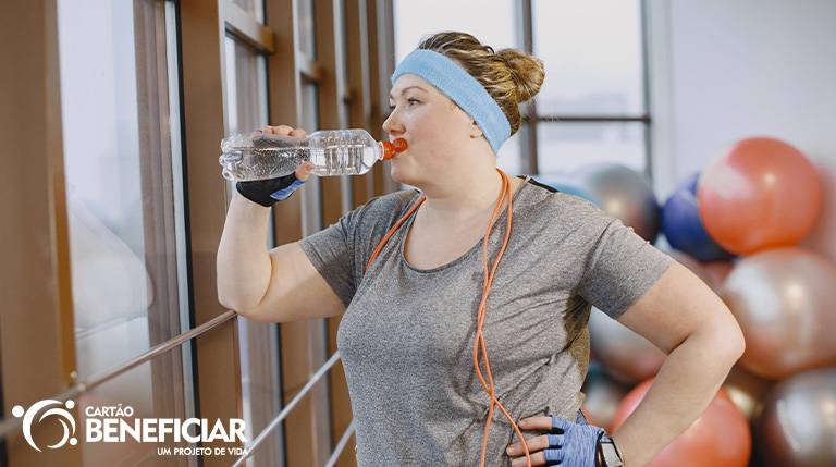 Mulher um pouco acima do peso na academia, bebendo uma garrafa de água com a mão na cintura enquanto olha pela janela. Ela é branca, loira, usa uma blusa cinza, luvas de musculação e uma faixa azul no cabelo, que está preso. Ao fundo, vemos algumas bolas de pilates.