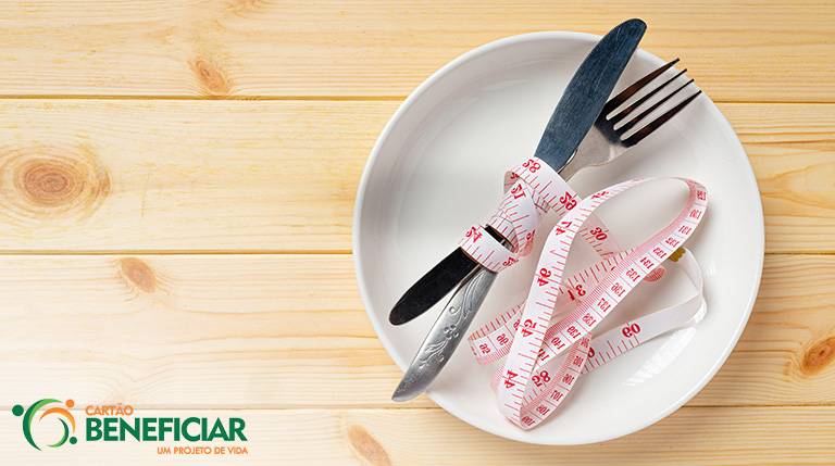 Um prato branco em cima de uma mesa de madeira marrom claro. Um garfo e uma faca estão cruzados em cima do prato e amarrados por uma fita métrica, representado a restrição alimentar, inimiga de um emagrecimento saudável.