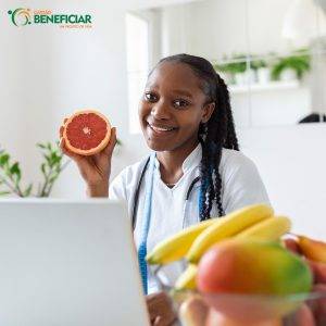 Nutricionista negra, com o cabelo trançado e usando um jaleco branco segura uma laranja. Em primeiro plano, estão algumas frutas indicadas para um emagrecimento saudável, como banana e manga, desfocadas. Ela está na frente de um notebook.