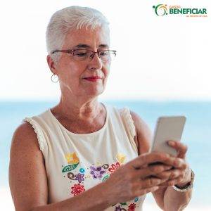 Mulher idosa, de cabelos brancos e curtos, olhando o celular com o braço esticado, um hábito comum de quem está com a vista cansada.