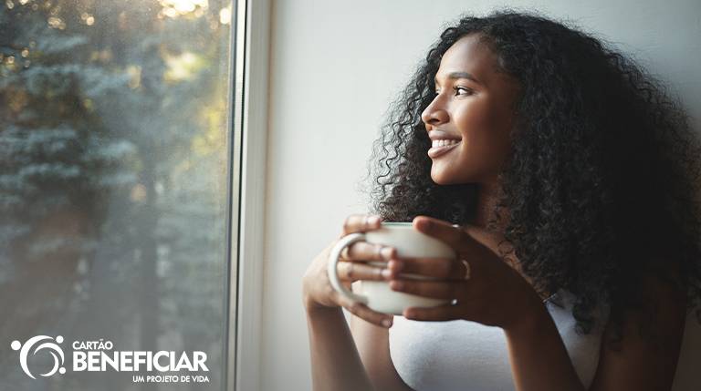 Mulher negra, de cabelos soltos e volumosos, usando uma blusa branca, sorri enquanto olha por uma janela. Ela segura uma caneca branca com as duas mãos e a paisagem que ela contempla é de uma floresta. A adesão ao tratamento devolve a qualidade de vida das mulheres com endometriose.