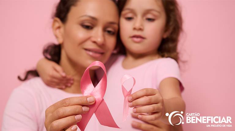 Mãe e filha abraçadas segurando uma fitinha rosa em primeiro plano representando a importância da prevenção do câncer de mama. O rosto delas está desfocado ao fundo e as mãos com a fitinha em primeiro plano.