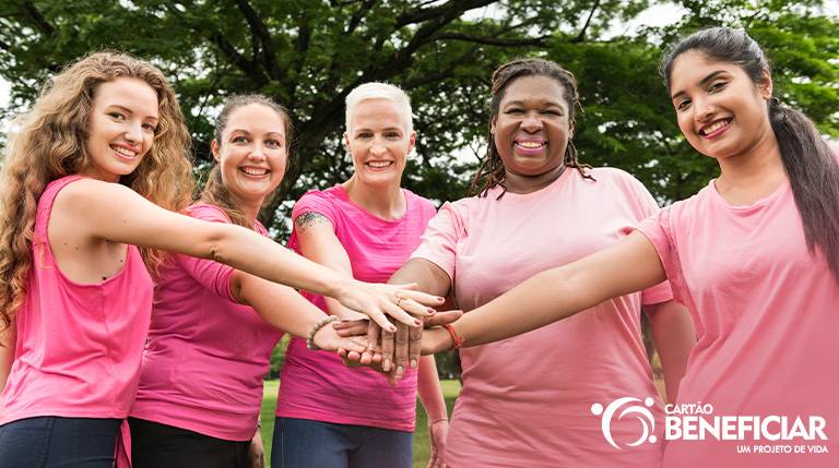 Cinco mulheres vestidas de rosa ao ar livre, com as mãos uma sobre a outra, em um gesto de força.