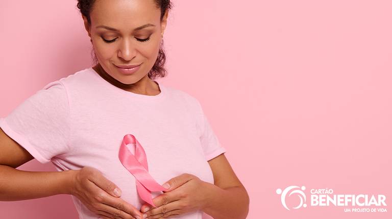 Mulher negra usando uma blusa rosa clara em fundo rosa mais escuro. Ela segura uma das mamas com as duas mãos e tem a fitinha rosa no peito.