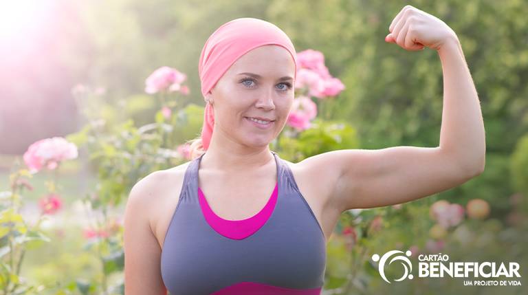 Mulher com lenço rosa ao ar livre, com um dos braços levantados simbolizando a força. A pose dela reforça a importância da prevenção do câncer de mama e do diagnóstico precoce para a sobrevivência ao câncer.