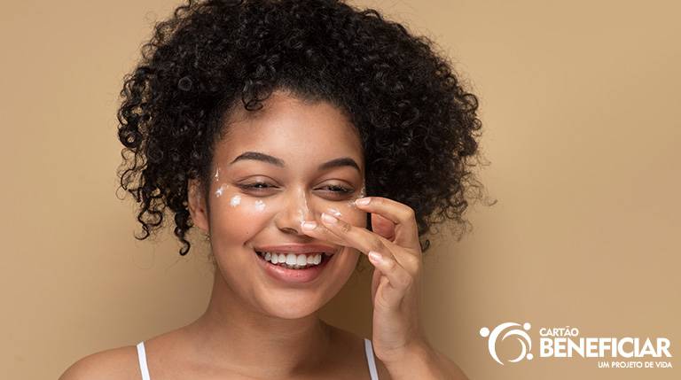 Mulher negra, de cabelos encaracolados, sorrindo e passando filtro solar no rosto