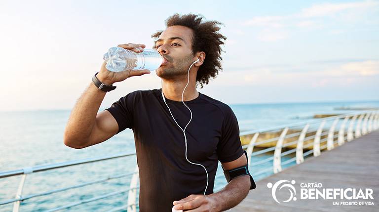 Homem negro praticando exercício na beira do mar, com fones de ouvido e tomando uma garrafa de água para se prevenir da desidratação, uma doença de verão