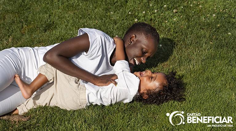 Mãe e criança deitadas na grama, sob o sol, brincando. Elas estão usando roupas claras, de tecido leve, e estão sorrindo