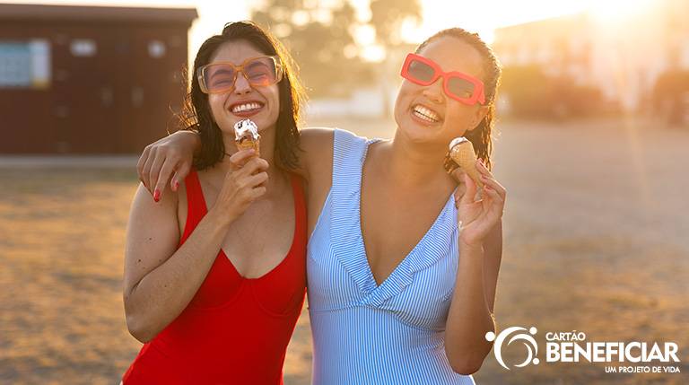 Duas amigas abraçadas na praia, ao entardecer, tomando sorvete e protegidas das doenças de verão. Elas são morenas, estão com o cabelo molhado e usando maiôs