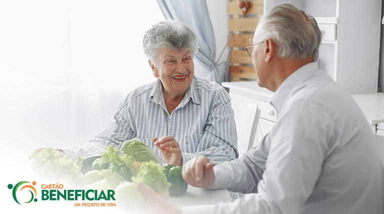 Uma mulher e um homem, ambos idosos, conversam sentados a uma mesa em que estão diversas hortaliças.