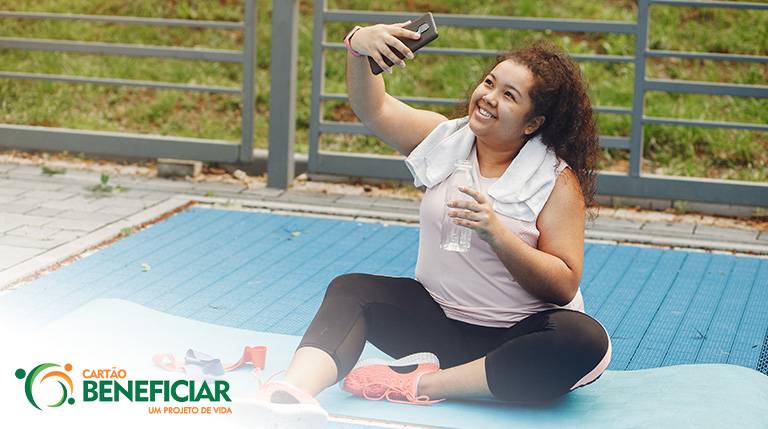 Menina tirando uma selfie com o celular. Ela está feliz, com roupas de ginástica, sentada em um colchonete, representando a atividade física como uma das melhores maneiras de lidar com todos os tipos de ansiedade.