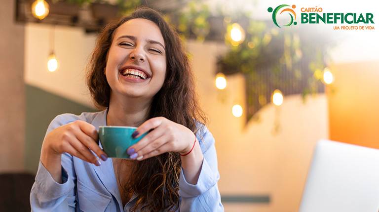 Mulher branca, de cabelos ondulados jogados de lado, sorrindo segurando uma xícara com as duas mãos, aproveitando um dos benefícios do café, que é a melhora do humor.