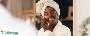 Mulher negra com uma toalha na cabeça e roupão limpando a pele com um algodão, parte fundamental de uma rotina de cuidados com a pele