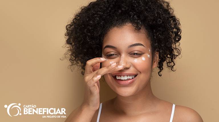 Mulher negra sorrindo, passando hidratante no rosto, uma das etapas de cuidados com a pele.