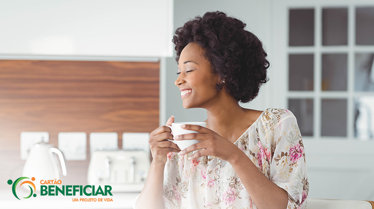 Mulher negra sorrindo de perfil segurando uma caneca de chá. Ela está em uma cozinha e usa uma blusa florida estampada.