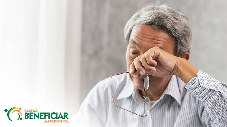 Homem asiático com uma camisa listrada coça os olhos com as costas da mão, em que segura seus óculos. Este é um sinal claro de que a pessoa precisa usar óculos ou ajustar o grau.