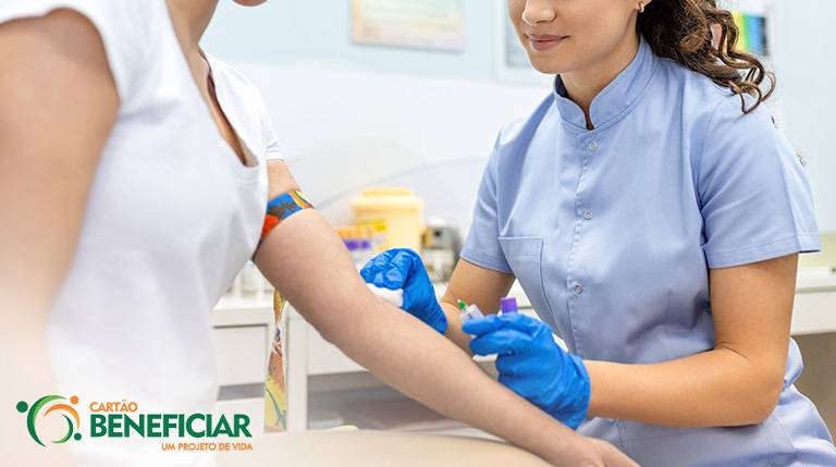 Enfermeira coletando sangue de paciente. A enfermeira é uma mulher branca, de cabelos encaracolados, veste azul e vemos apenas a metade inferior do seu rosto e suas mãos, com luvas azuis. Só vemos o braço da paciente e seu tronco. Ela é uma mulher branca vestida de branco. Elas estão em uma sala de coleta de exames.