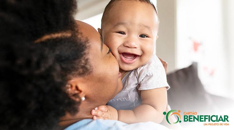 Bebê sorrindo feliz olhando diretamente para a câmera. Ele está no colo da mãe, uma mulher negra usando um brinco de pérola, e o rosto dela está de perfil, sorrindo, com o rosto apoiado no dele.