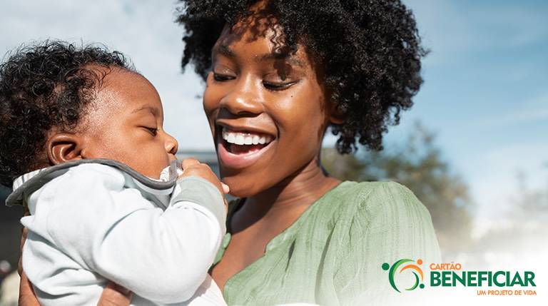 Uma mulher negra, com um sorriso largo, carrega seu bebê, que parece já ter aproximadamente um aninho. Ela está olhando para ele muito feliz e ele está com a mãozinha na boca.