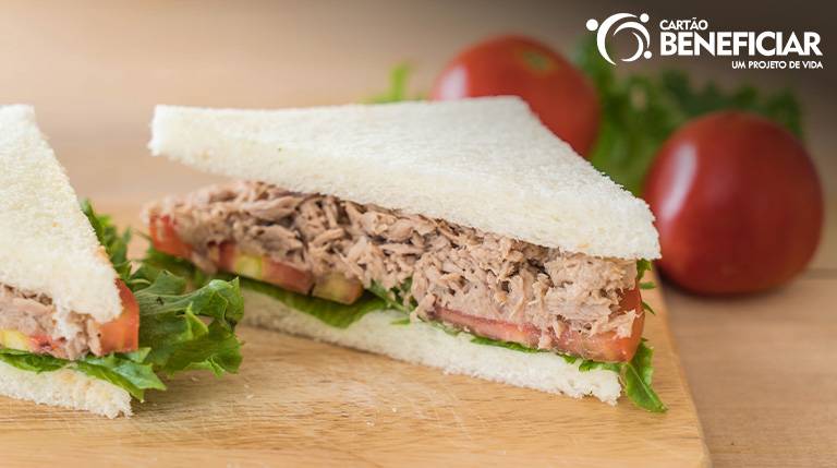 Imagem ilustrativa de um sanduíche natural de atum em pão de forma. O lanche tem alface, tomate, atum e está cortado em formato triangular.