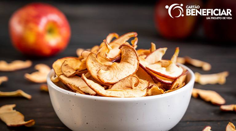 Imagem ilustrativa de chips de maçã. Eles estão sobre uma mesa escura, dentro de uma tigela branca.