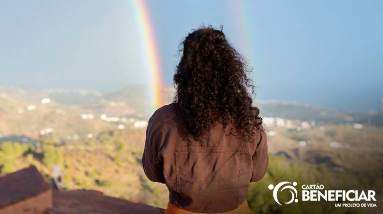 Mulher de costas em pé, em um lugar elevado, olhando para um arco-íris que se forma no horizonte.