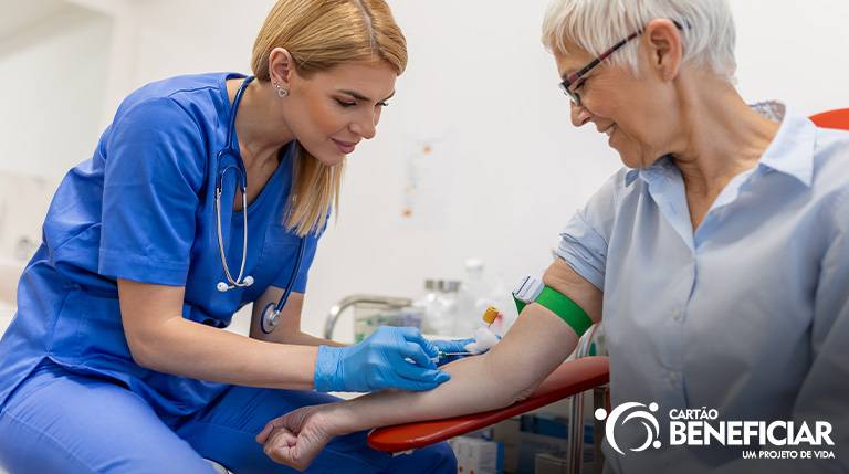 Uma enfermeira loira, vestida de azul e com um estetoscópio no pescoço faz a coleta de sangue de uma paciente idosa. Ela tem um garrote no braço e está sorrindo durante o exame de sangue.