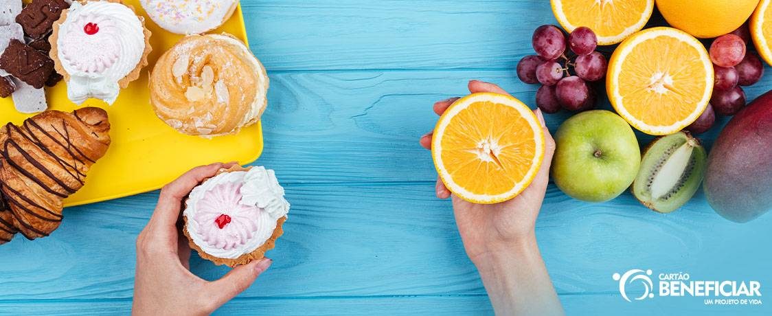 A imagem mostra só as mãos de uma pessoa segurando um cupcake e meia laranja. Do lado do cupcake, tem doces e massas. Do lado da laranja, tem frutas. O objetivo é representar o equilíbrio necessário para ter uma alimentação saudável.