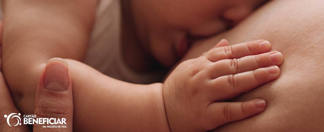 Bebê sendo amamentado no peito e recebendo todos os benefícios da amamentação. Em primeiro plano, vemos sua mãozinha apoiada no seio da mão e também o polegar da mãe segurando seu bracinho. Em segundo plano, desfocado, está seu rostinho sendo amamentado.