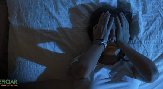 Mulher deitada na cama, em um quarto escuro, com as duas mãos sobre o rosto, em um gesto que indica o desespero de não conseguir dormir.