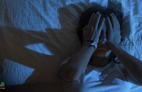 Mulher deitada na cama, em um quarto escuro, com as duas mãos sobre o rosto, em um gesto que indica o desespero de não conseguir dormir.
