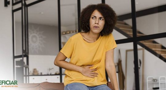 Mulher negra, de cabelos crespos soltos, vestindo uma blusa amarela e calça cinza, com uma expressão de dor e pressionando a região do estômago em decorrência dos desconfortos causados pelo refluxo.