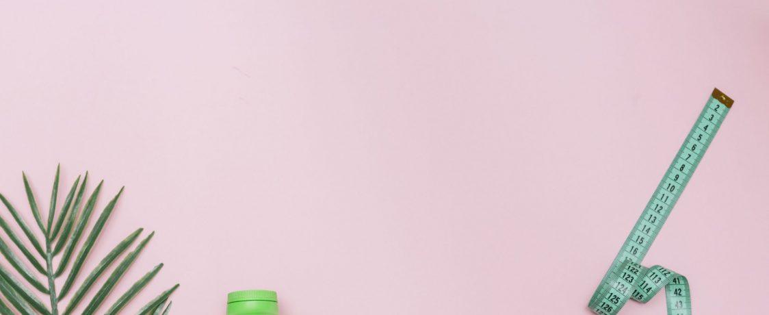 Pedaço de laranja, copo de água, tênis e fita métrica lado a lado em fundo rosa, representando o que é necessário para perder peso.
